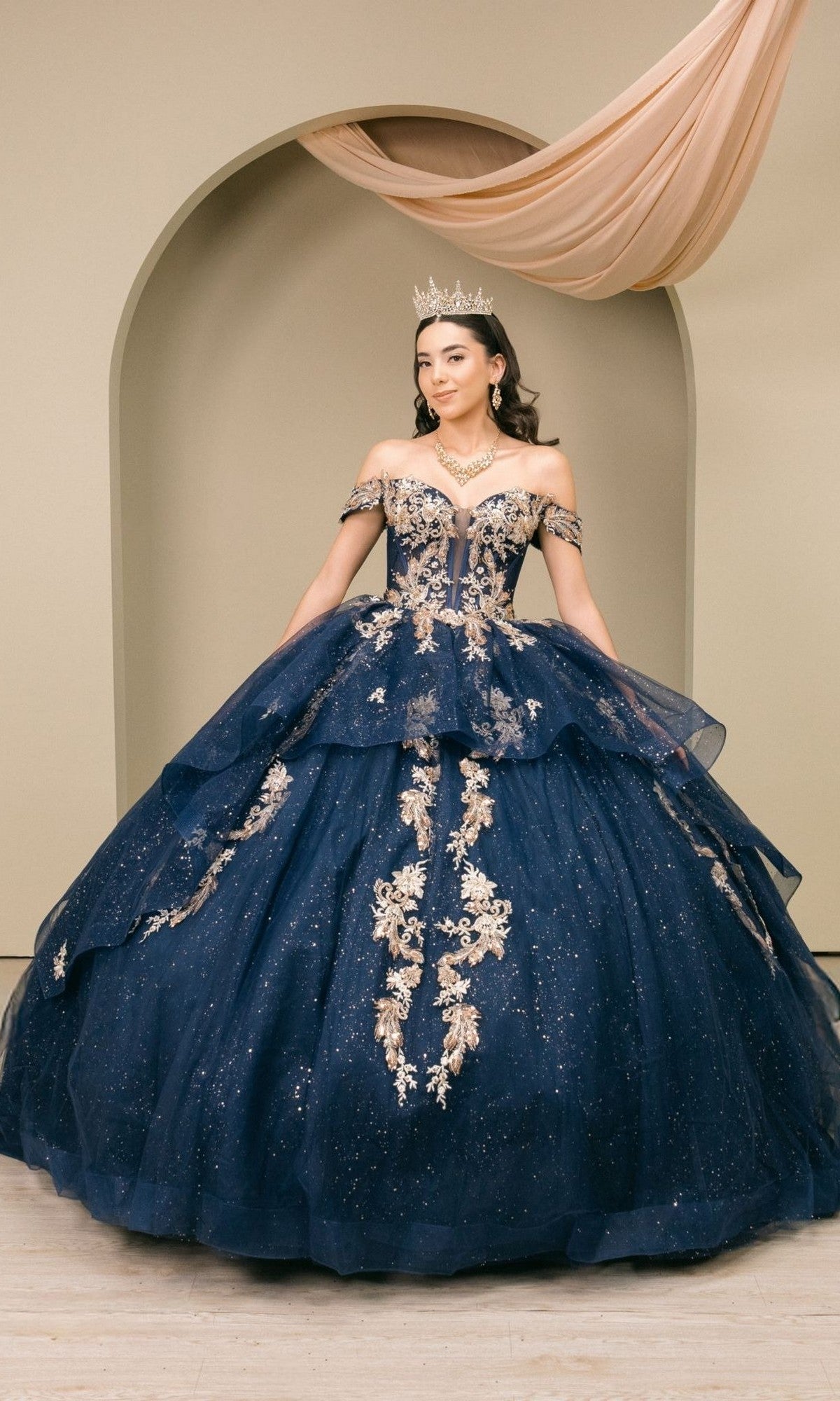 A woman wearing the Quinceanera Dress 8390JQ by Cinderella Couture in navy, featuring intricate gold embroidery, a crown, and sparkling jewelry, stands gracefully before a beige arch backdrop draped with elegant fabric.